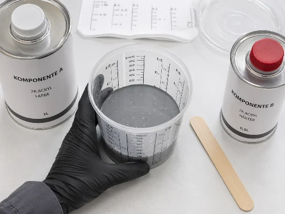 Draufsicht auf einen sauberen Mischtisch zum Anmischen von 2K-Acryllack. In der Mitte steht ein transparenter Mischbecher mit Skala und grauer Lackmischung. Eine Hand mit schwarzem Schutzhandschuh hält den Becher fest. Links daneben steht eine Dose mit der Beschriftung „Komponente A“, darunter „2K Acryl Farbe“ und „1L“. Rechts steht eine kleinere Dose mit rotem Deckel und der Beschriftung „Komponente B“, darunter „2K Acryl Härter“ und „0,5L“. Neben dem Mischbecher liegt ein Holzrührspatel. Im Hintergrund sind weitere Mischhilfen und ein transparenter Deckel zu sehen. Das Bild zeigt übersichtlich die wichtigsten Materialien zum korrekten Mischen von 2K-Acryllack und Härter.