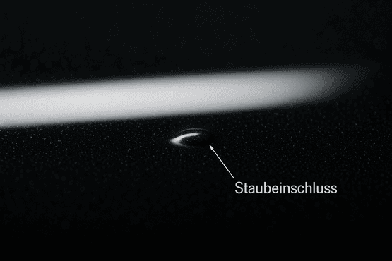Makrofoto als Beispiel für Staubeinschlüsse. Es zeigt einen einzelnen Defekt auf schwarzem Hochglanzlack, auf den ein Pfeil mit der deutschen Aufschrift 'Staubeinschluss' zeigt.