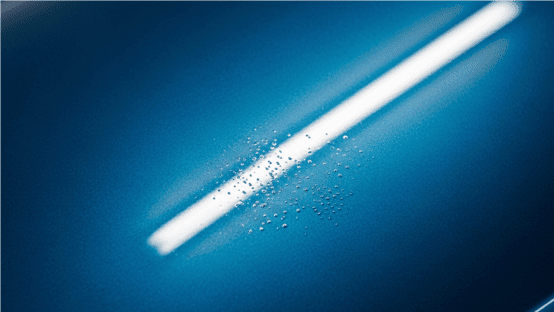 Nahaufnahme einer blau-metallic Autolackierung mit dem Lackierfehler Kocher. Eine helle Lichtreflexion hebt die feinen Bläschen und die unebene Oberflächenstruktur hervor.
