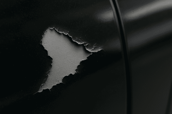 Nahaufnahme von Haftungsstörungen: Schwarzer Autolack platzt von der grauen Grundierung ab und zeigt einen typischen Lackschaden.