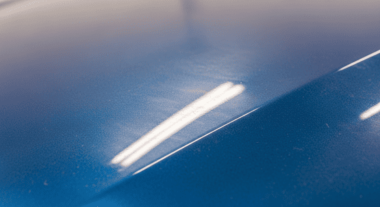 Eine extreme Nahaufnahme von blauem Autolack mit einem schweren Lackierfehler, der als Vermattung bekannt ist. Die Oberfläche ist komplett stumpf, hat keinerlei Glanz und die Reflexion einer Werkstattlampe ist verschwommen und diffus statt scharf und klar.