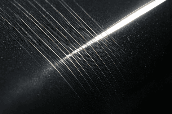 Eine extreme Nahaufnahme einer dunkelgrauen, metallischen Autolackoberfläche, die von einer hellen Lichtquelle angestrahlt wird. Deutlich sichtbar sind feine, parallele Schleifriefen, die unter der glatten, glänzenden Klarlackschicht eingeschlossen sind und das Licht reflektieren.