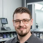 Quadratisches Porträt von Marcel Prinz von Lackundzubehoer.de. Der Experte blickt freundlich in die Kamera, trägt eine Brille und ein graues Poloshirt. Im Hintergrund ist unscharf die professionelle Ausstattung einer Lackierwerkstatt zu erkennen.