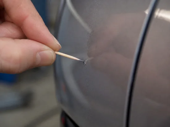 Nahaufnahme einer grauen Autotür mit kleinem Lackschaden. Eine Hand führt einen Zahnstocher an die beschädigte Stelle, um Lack oder Reparaturmaterial gezielt in den Steinschlag einzubringen. Rechts ist eine Türfuge zu sehen, der Hintergrund ist unscharf.