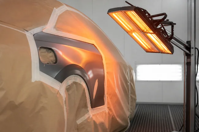 Ein teilweise abgedecktes Fahrzeug steht in einer Lackierkabine. Braunes Abdeckpapier und transparente Folie schützen die Karosserie, während rechts ein eingeschalteter Infrarot-Trockner orange leuchtet und Wärme auf den lackierten Bereich richtet.
